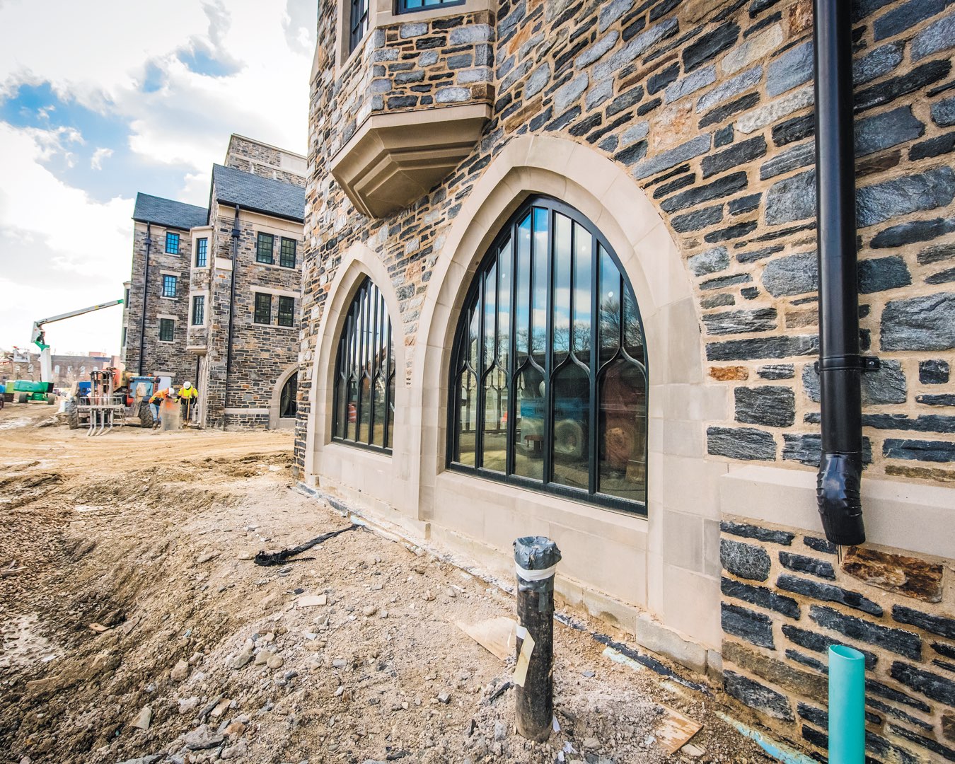 Black gothic arched windows on stone facade of The Commons residence halls, with construction crew in the distance on site 