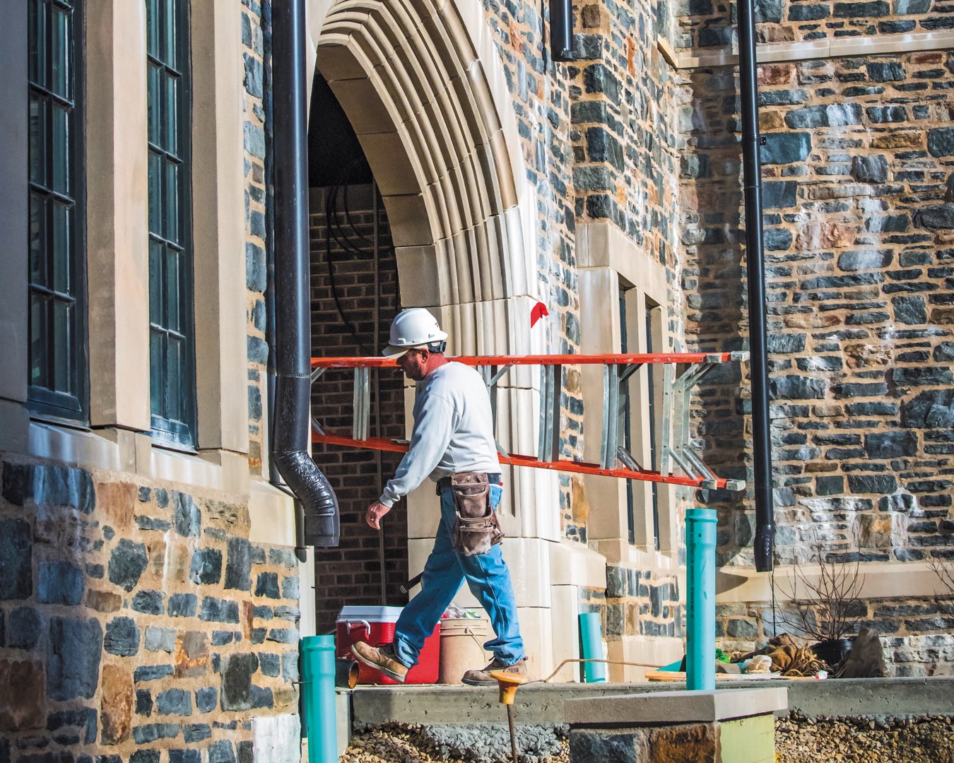 A construction worker carries a ladder under one of the stone arches of The Commons residence halls