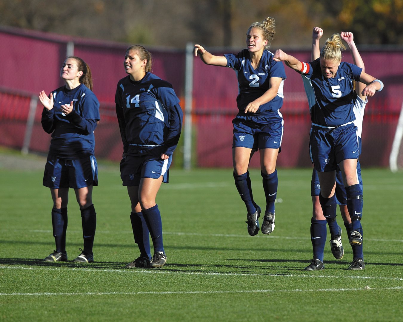 Five Villanova Women’s Soccer players in uniform on the field clapping, cheering and jumping in the air
