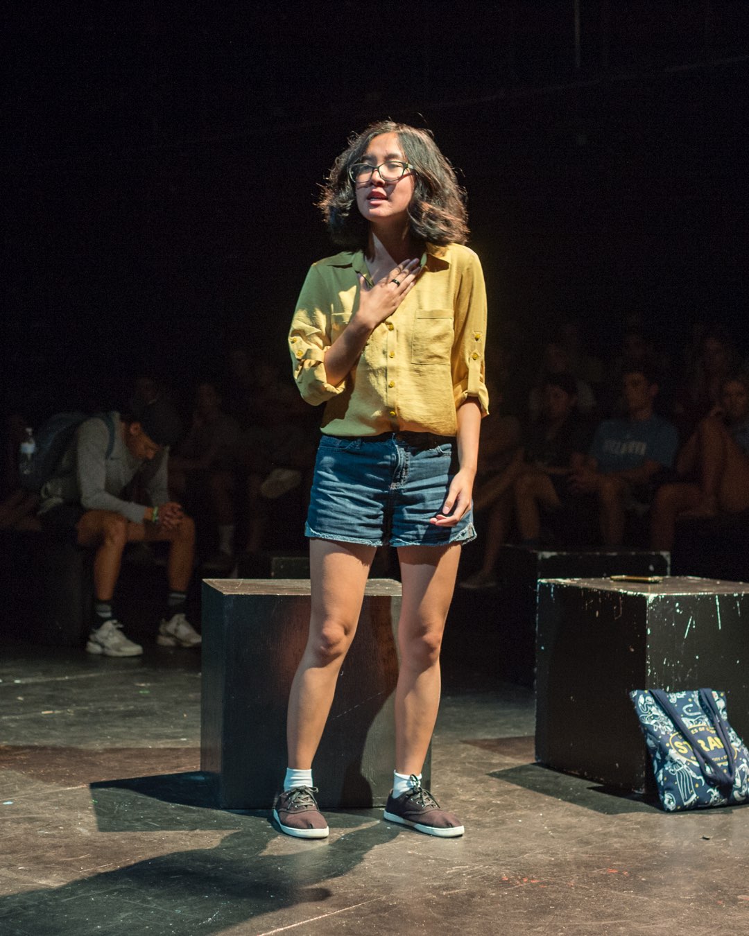 A female student in glasses with a yellow shirt and denim shorts, speaking with her hand against her chest