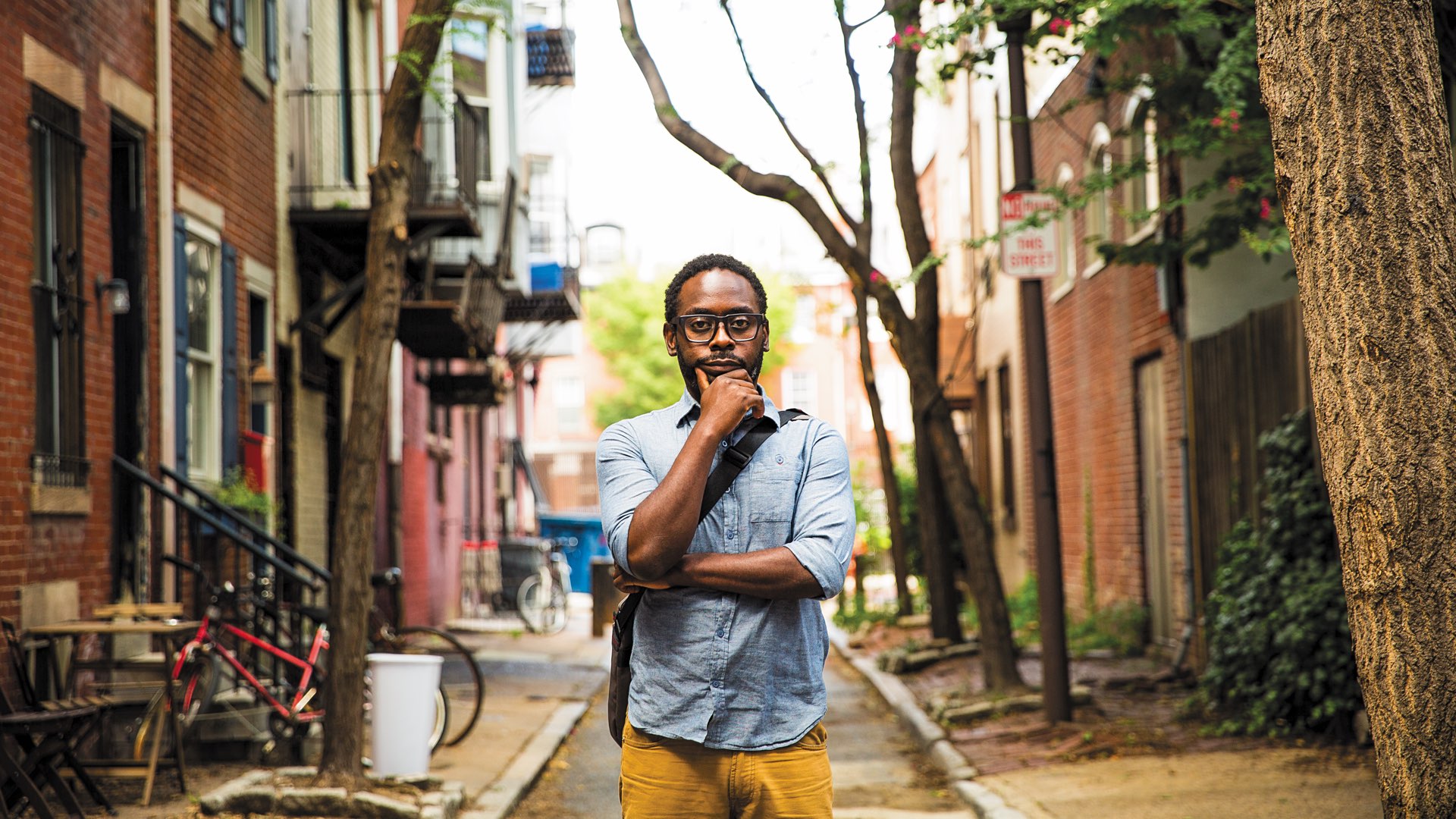 Professor James Ijames in a blue button down and mustard-colored jeans in the middle of a tree-lined street in Philadelphia