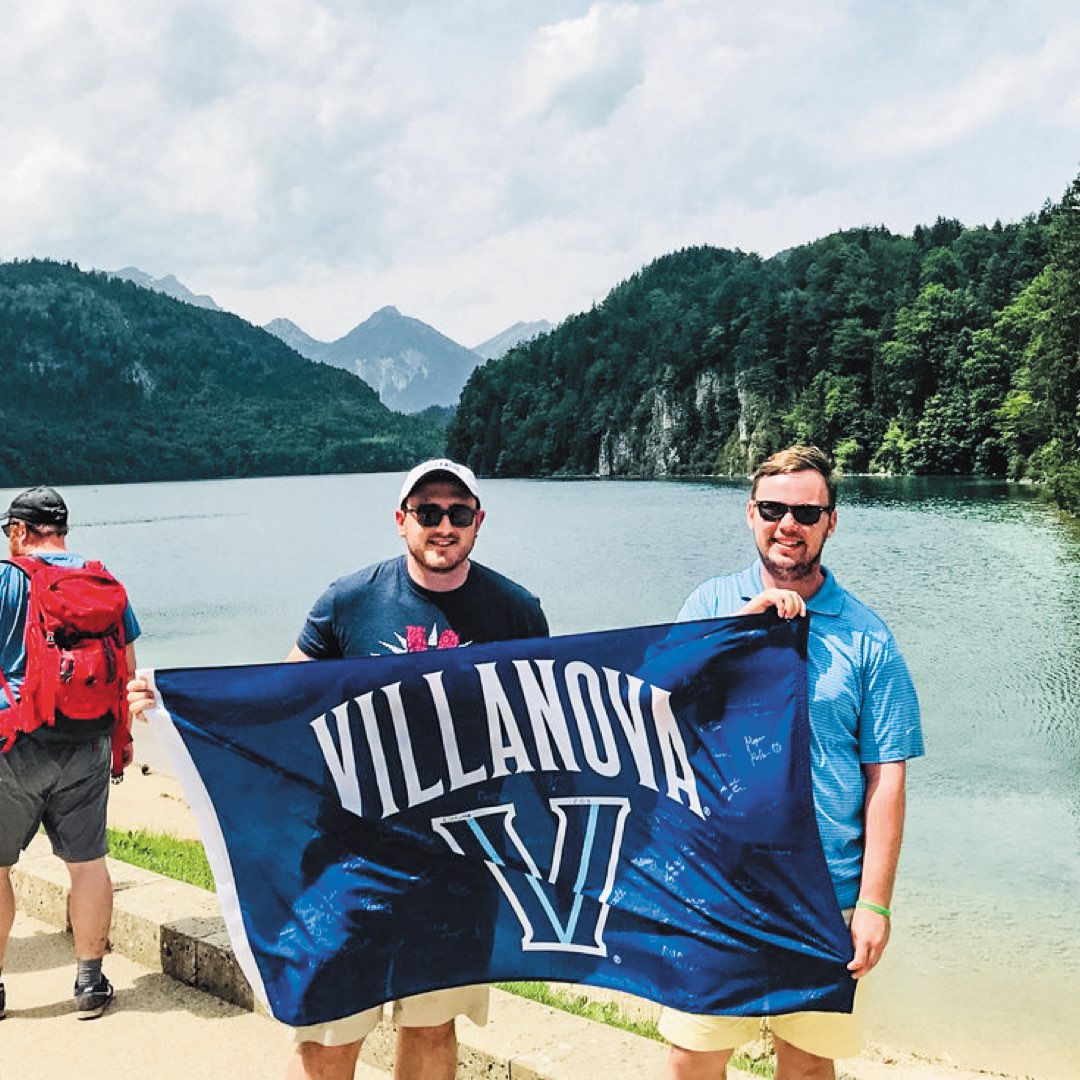 Two Villanova male alumni holding a Villanova banner with Lake Alpsee, Germany in the background