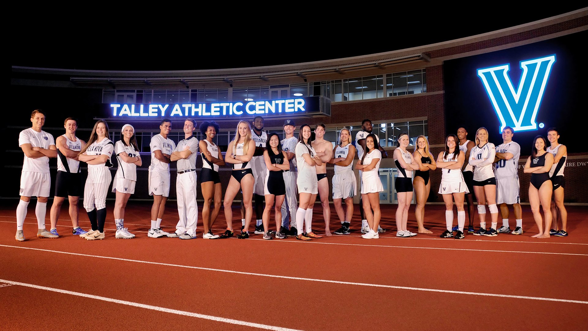 Villanova student-athletes stand in front of the Andrew J. Talley Athletic Center, the blue signs glowing at night.