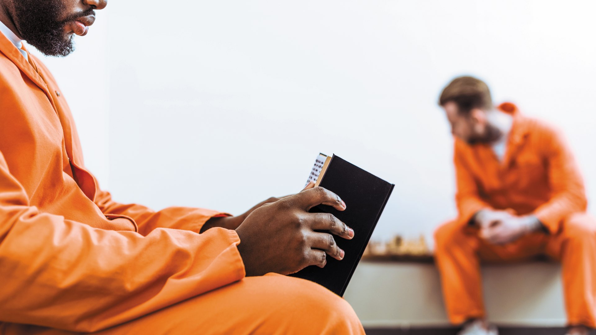 A prison inmate wearing an orange jumpsuit reading an open book with another inmate in the background 