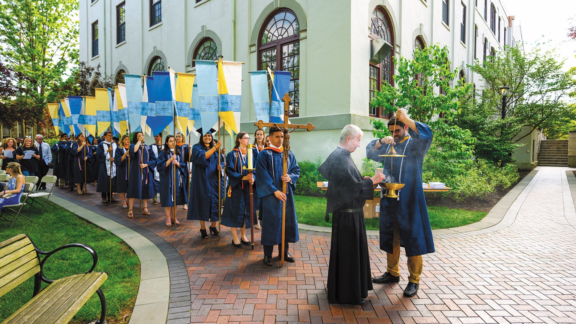 Students in graduation regalia holding banners line up for the Baccalaureate Mass procession on campus