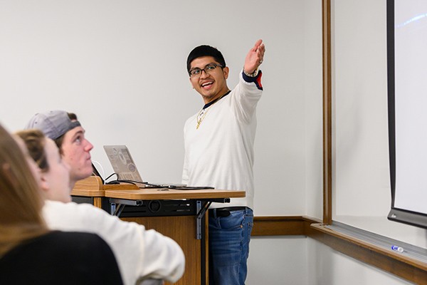 A student raises his arm to point to a projection while he presents to other students in class.