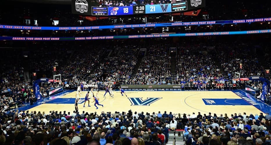 Wide shot of a basketball arena, filled with fans; the court has a Villanova "V" in the middle