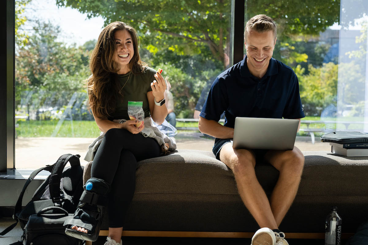 Two students sitting on a bench, smiling and looking at a computer.