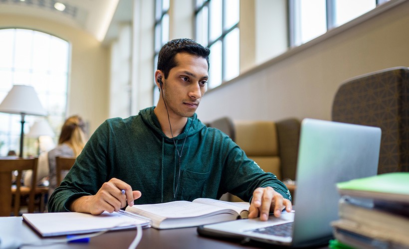 A student wearing earphones works at a laptop in the library reading room.