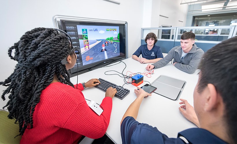 Four students sit around a table with a large screen showing a car racing video game working on a project to measure focus and attention levels in players with ADHD.