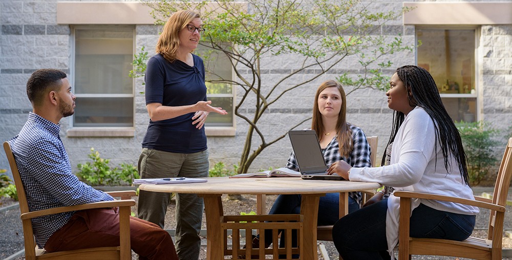 Faculty member talking with graduate students seated at an outdoor table.