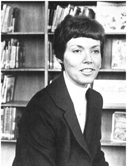 A black and white photo of a white woman with short hair in a suit,  near a bookcase