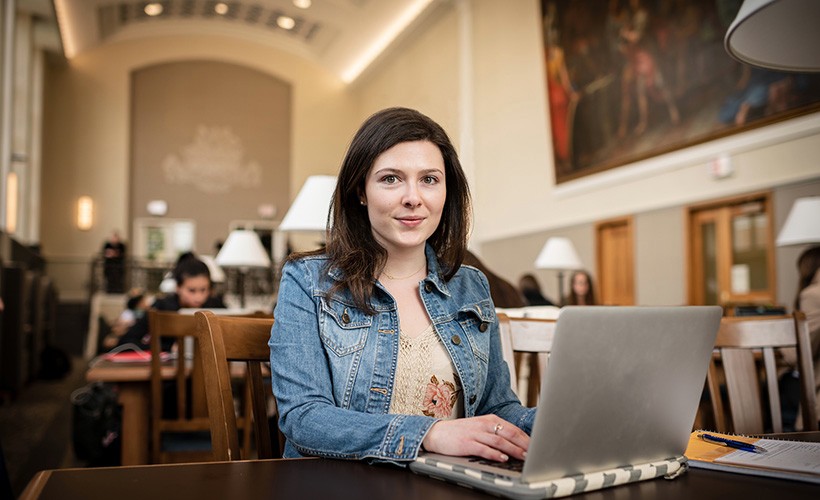 Student sits at a desk in the library with her computer open in front of her.