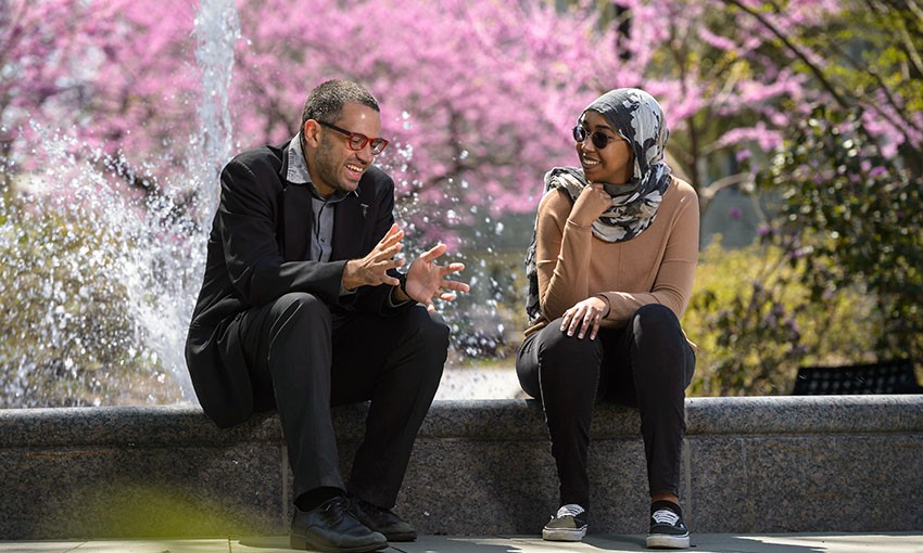 Theology professor talking to a student in front of a fountain on a spring day