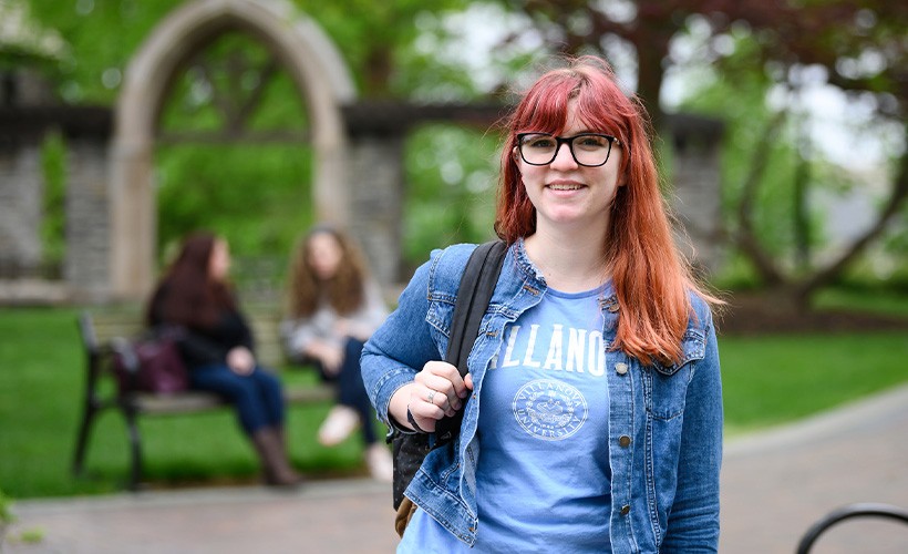 A student stands on a pathway on Villanova campus with two other students sitting on a bench in the background.