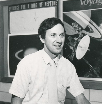 Dr. Guinan sits in front of his research team's poster displaying evidence for a ring around Neptune in 1982. A poster of the Voyager missions hangs adjacent, foreshadowing Voyager 2's photographic confirmation of the the planet's ring system in 1989. 