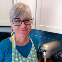 Female chef with grey hair and glasses stands in a kitchen near a mixer