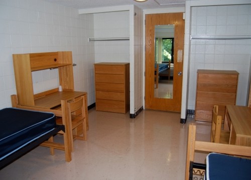 View of two beds, two desks and two open closets with dressers in a double room.