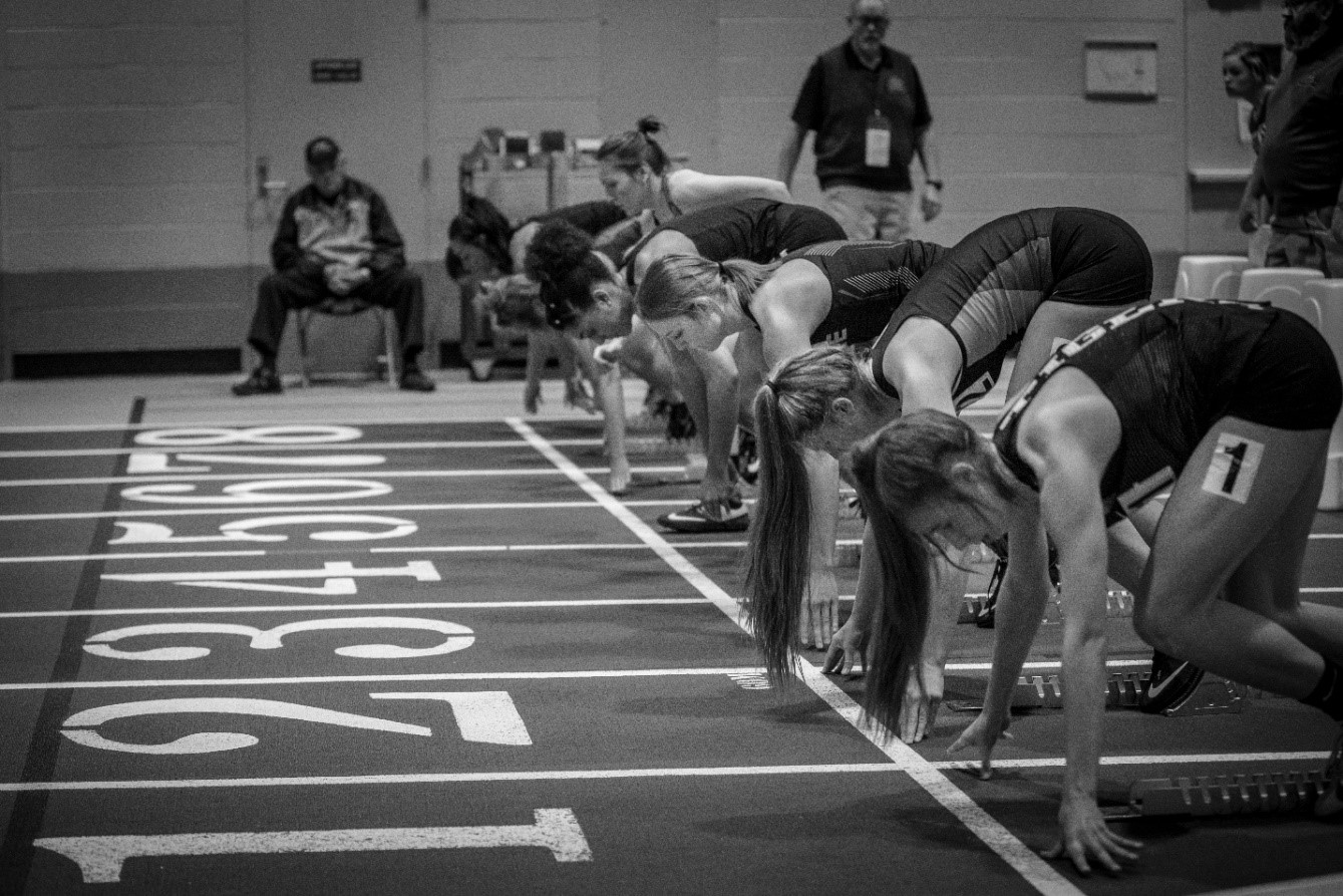 black and white image of female track athletics in position at starting line of indoor track meet