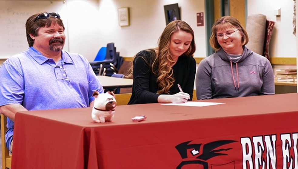female student athlete signing letter of intent seated at a table with a red table cloth with her parents on either side 