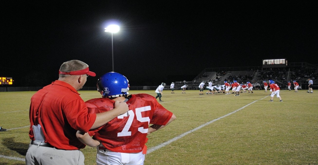 High school football coach and player stand shoulder to shoulder in red jerseys, staring at a field of other football players before player heads back into game