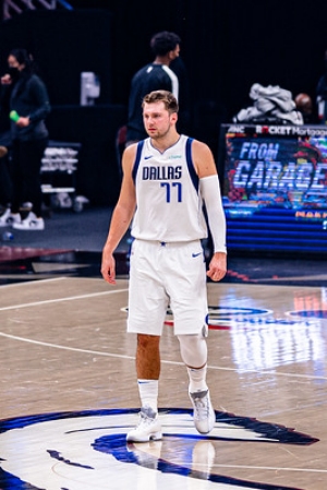 Photo of basketball player Luka Donic showing him walking across a basketball court wearing a white jersey that says Dallas 77 in blue text