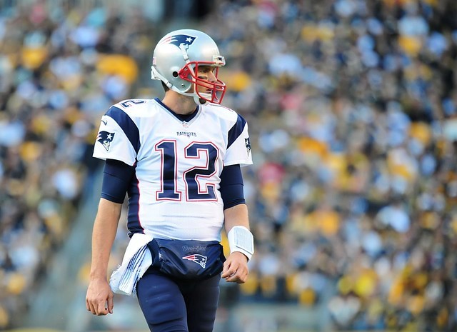 NFL Player Tom Brady Standing on field wearing white number 12 New England Patriots Jersey with blurred crowd in background