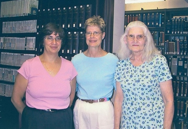 Photo of three former library staff members standing in front of library bookcases. From left to right: Regina Kozul, Mary Cornelius, and Maura Buri