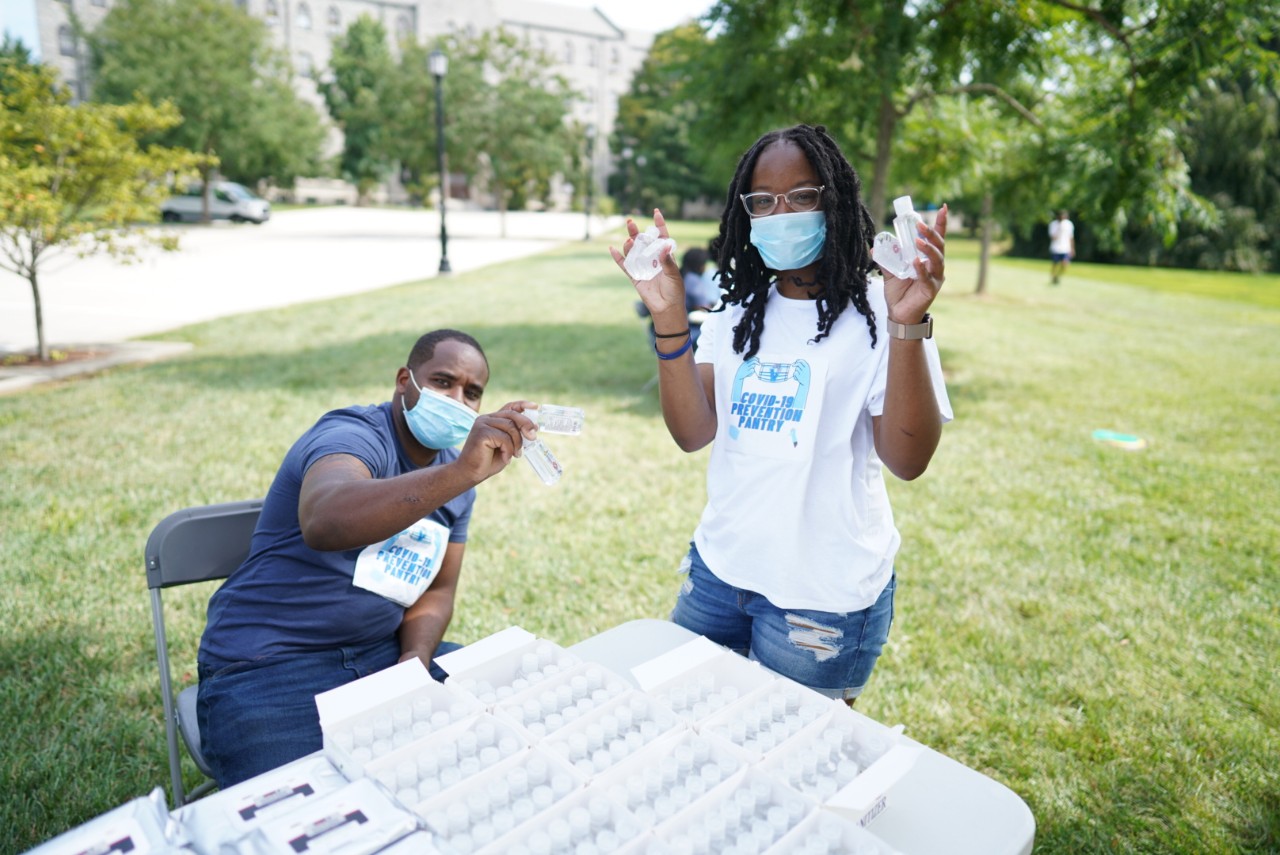 An image of Jasmine Mays and Tyshaan Williams. Both are standing arounf a table with COVID prevention kids on them, wearing masks and COVID prevention Pantry t-shirts.