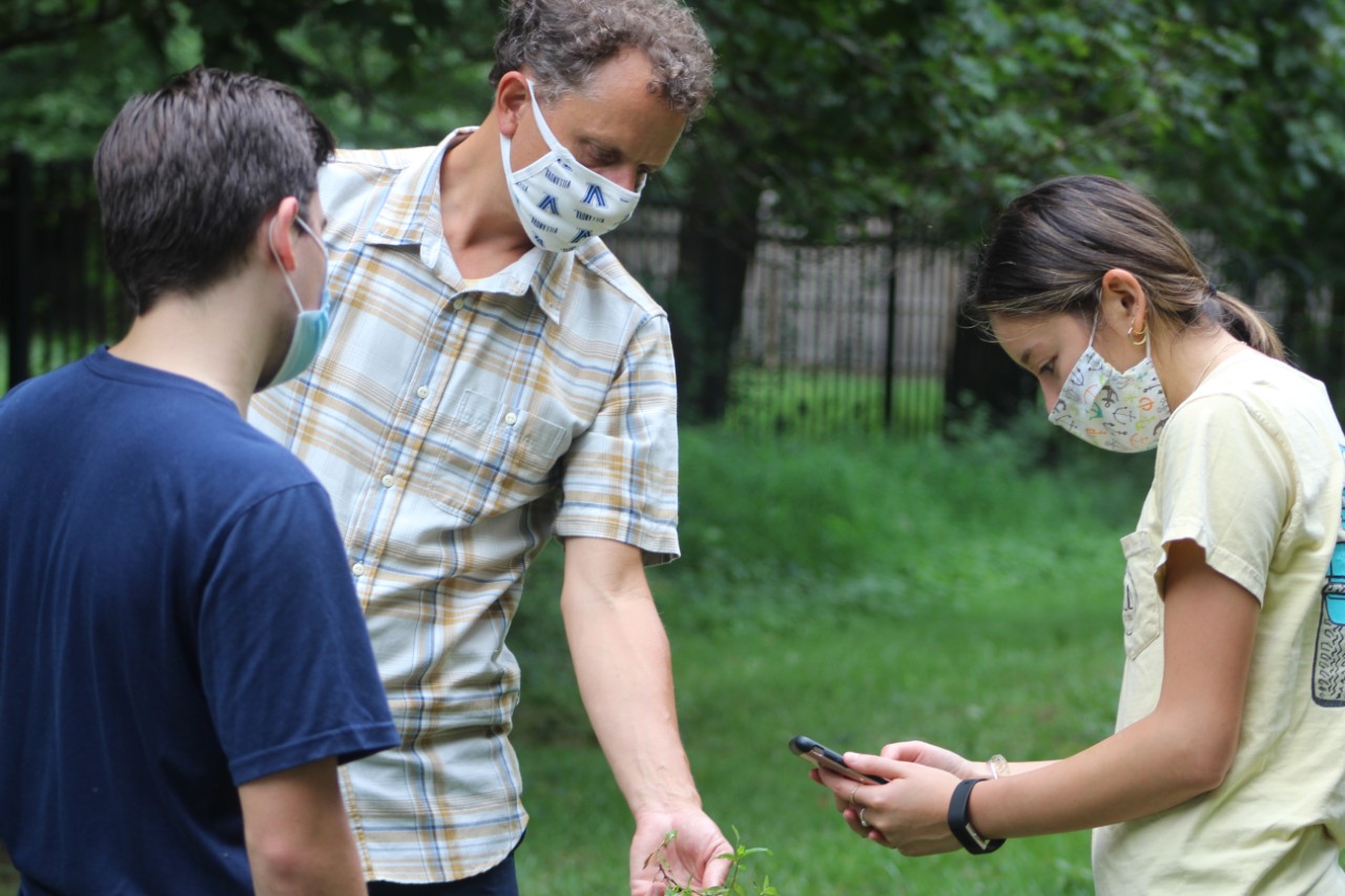 Adam Langley is in the center, wearing a cloth medical mask and a yellow checkered shirt. He holds out a tuft of grass, that the student to his left is taking a photo of with her phone. On Adam's right is a nother student looking on.
