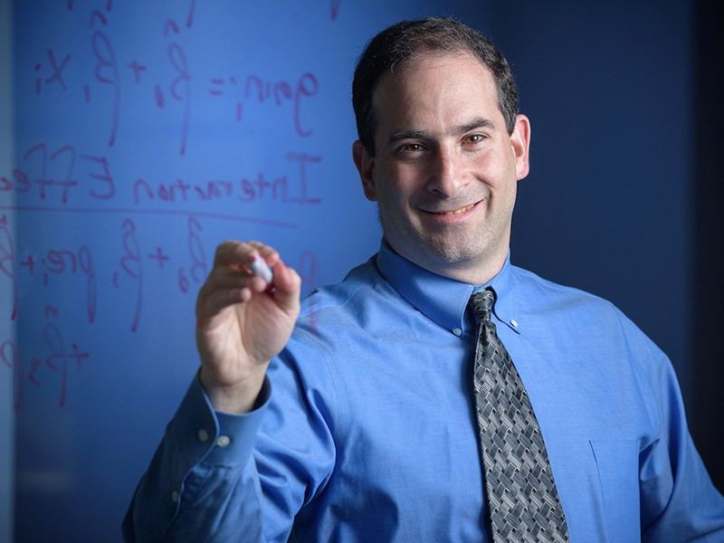 Photo of Dr. Posner from the waist up, standing in front of a white board with some figures on it. He is wearing a blue shirt with a pattenred grey tie. He is smiling, and holds a white0board marker in his hand.