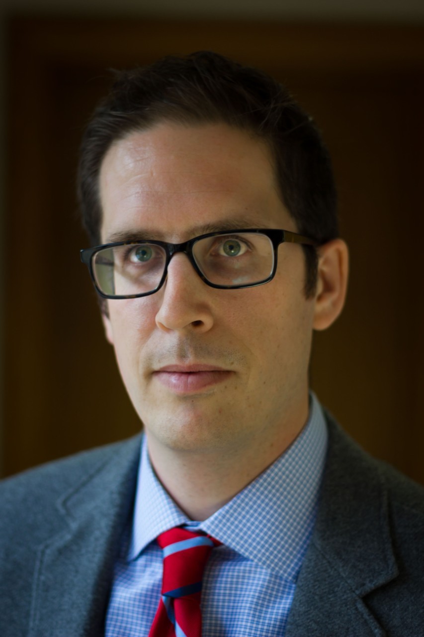 A photo of  Dr. McGuire from the shoulders up. He is a white man with short brown hair and wears black-framed glasses and a blue checkered shirt, red tie and gray suit jacket.