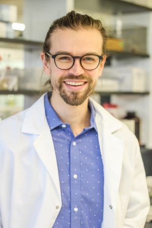Photo of Dr. Matthew O'Reilly standing in his lab, wearing a white lab coat and glasses. He is smiling.