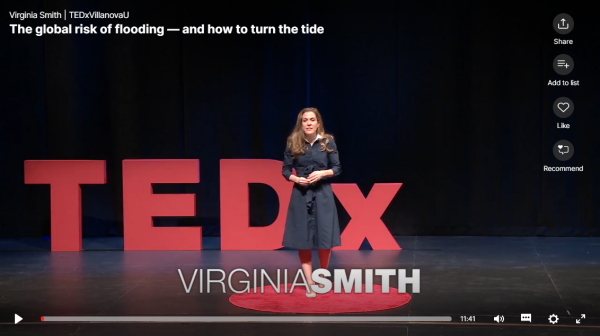 Screen shot of a video of Virginia Smith standing on a red capret on a stage with a TEDx sign behind her. 