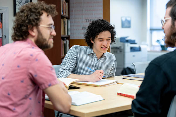 Male student working at a desk with two peers chatting in foreground