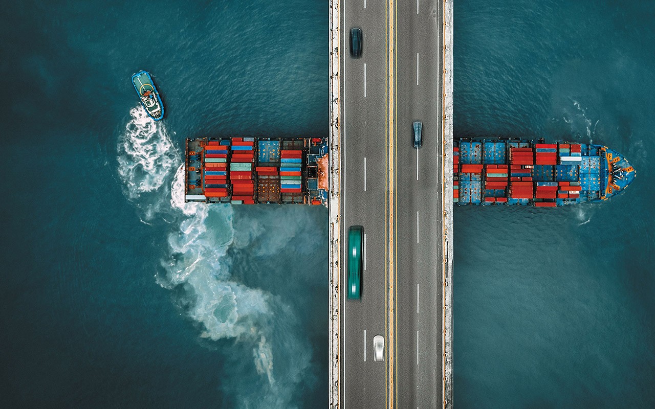 aerial view of a cargo ship loaded with containers passing under a bridge