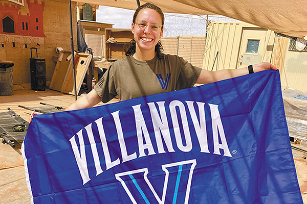 Lt. Cardon Furry standing outside and smiling on a US Army base in Syria holding a Villanova flag