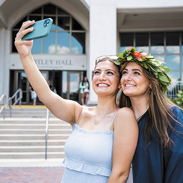 two smiling young alumni pose for a selfie in front of Bartley Hall