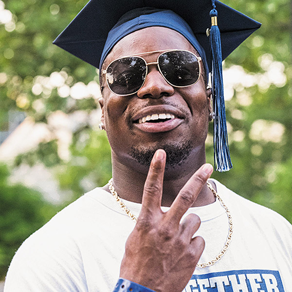 a young alumni wearing sunglasses and a graduation gives a Vs up to the camera