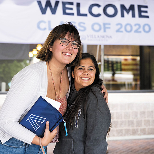 two young alumni stand close together posing for a photo in front of a Welcome Class of 2020 banner