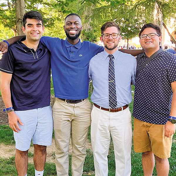 four young alumni smile at the camera while standing side-by-side on a sunny day