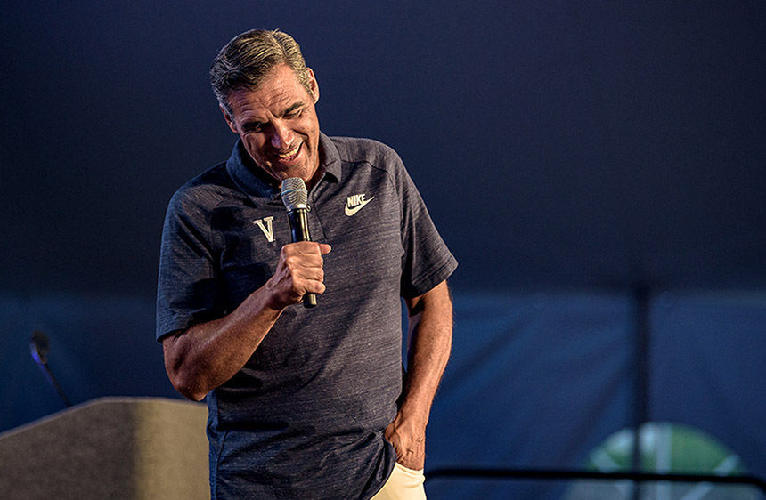 Jay Wright dressed in a Nike Villanova grey polo looks down as he holds a microphone laughing