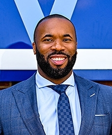  a smiling Kyle Neptune dressed in a blue suit and tie standing against a brick wall