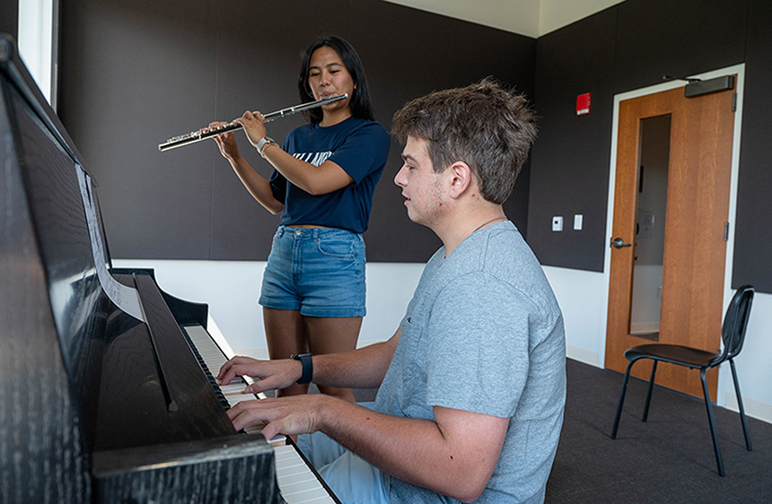 a female student stands and plays the flute while a male student is seated and plays the piano