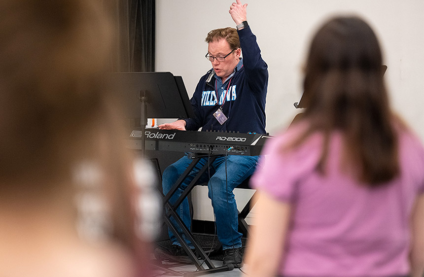 an instructor in a Villanova sweatshirt plays the keyboard as he directs singers 