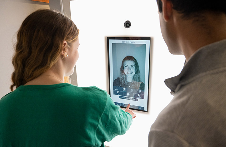 two people stand in a self-service photo booth and look at a headshot on a screen