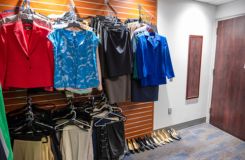 a wall is filled with hangers of business attire and on the carpeted floor is a row of women's pumps