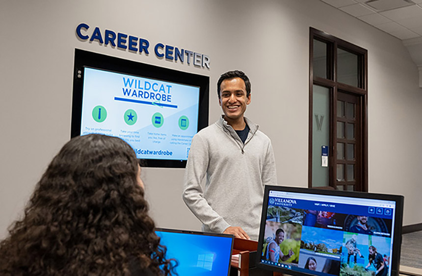 a smiling man stands in the Career Center lobby looking at a person seated before two computer monitors
