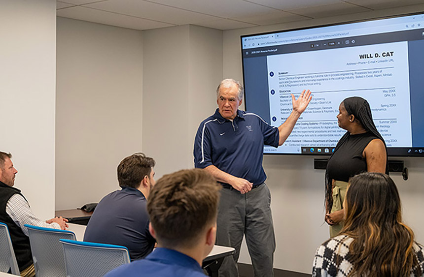 an older man wearing a Nike Villanova blue polo and grey pants stands in a classroom in front of a smartboard projecting a resume for Will D. Cat