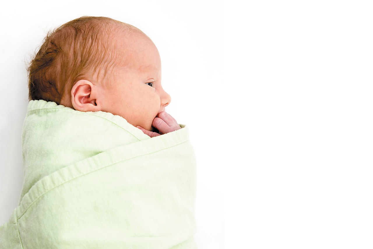 a newborn baby with their fist in mouth laying sideways swaddled in a light-colored blanket